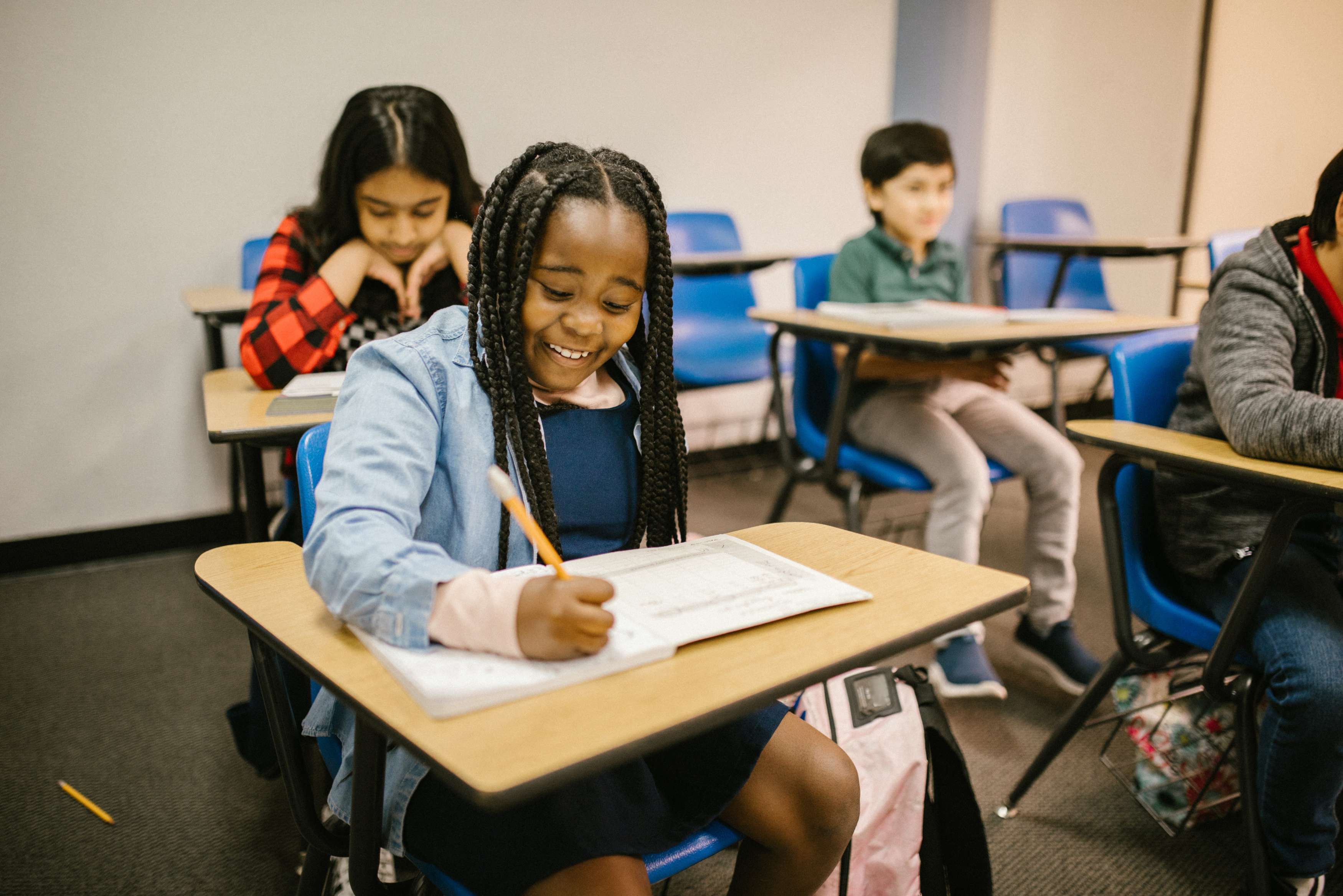 A smiling girl with braids writing in class, while other students focus on their work.