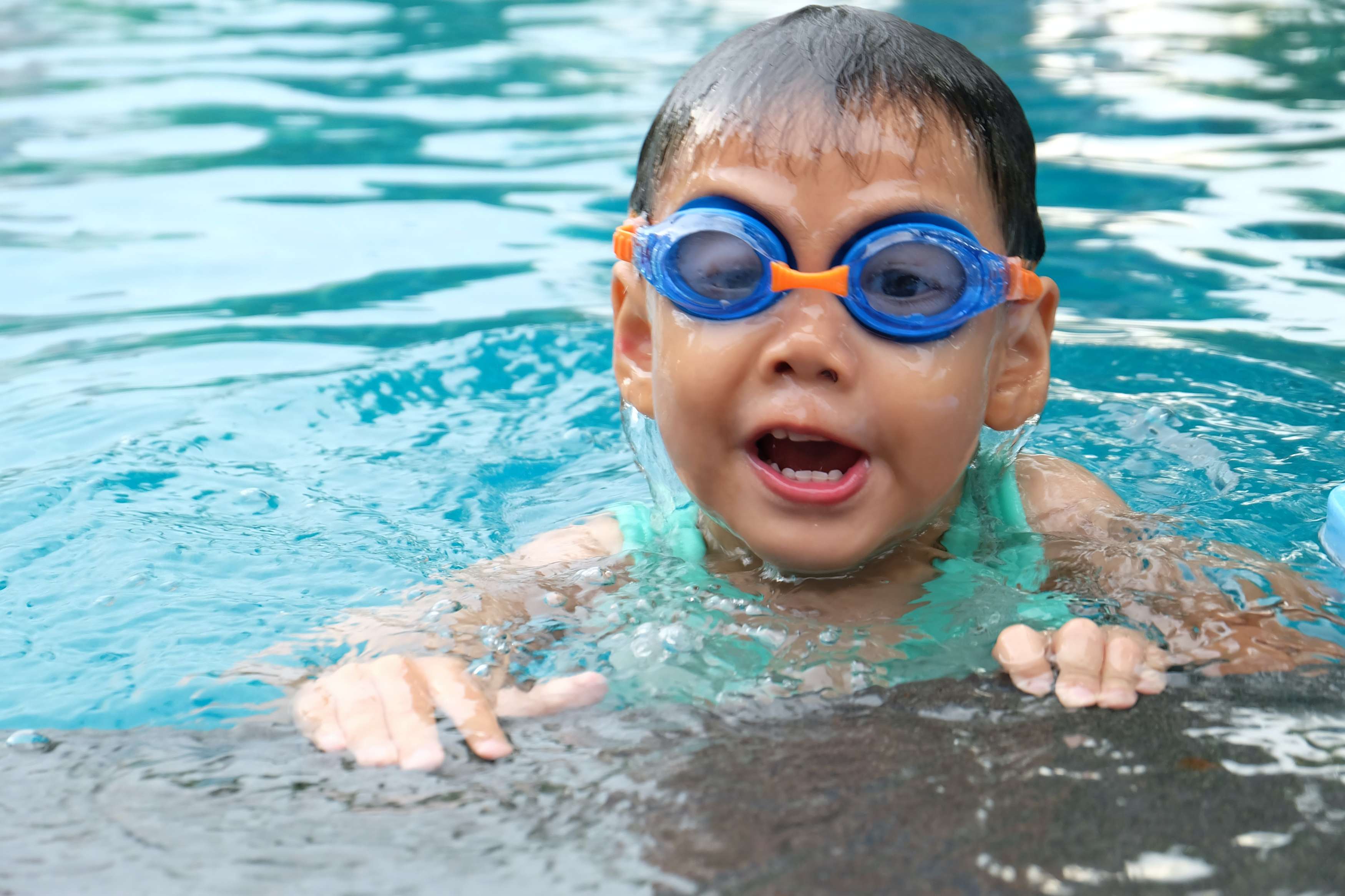 A young boy with swim goggles smiles while floating in a pool.